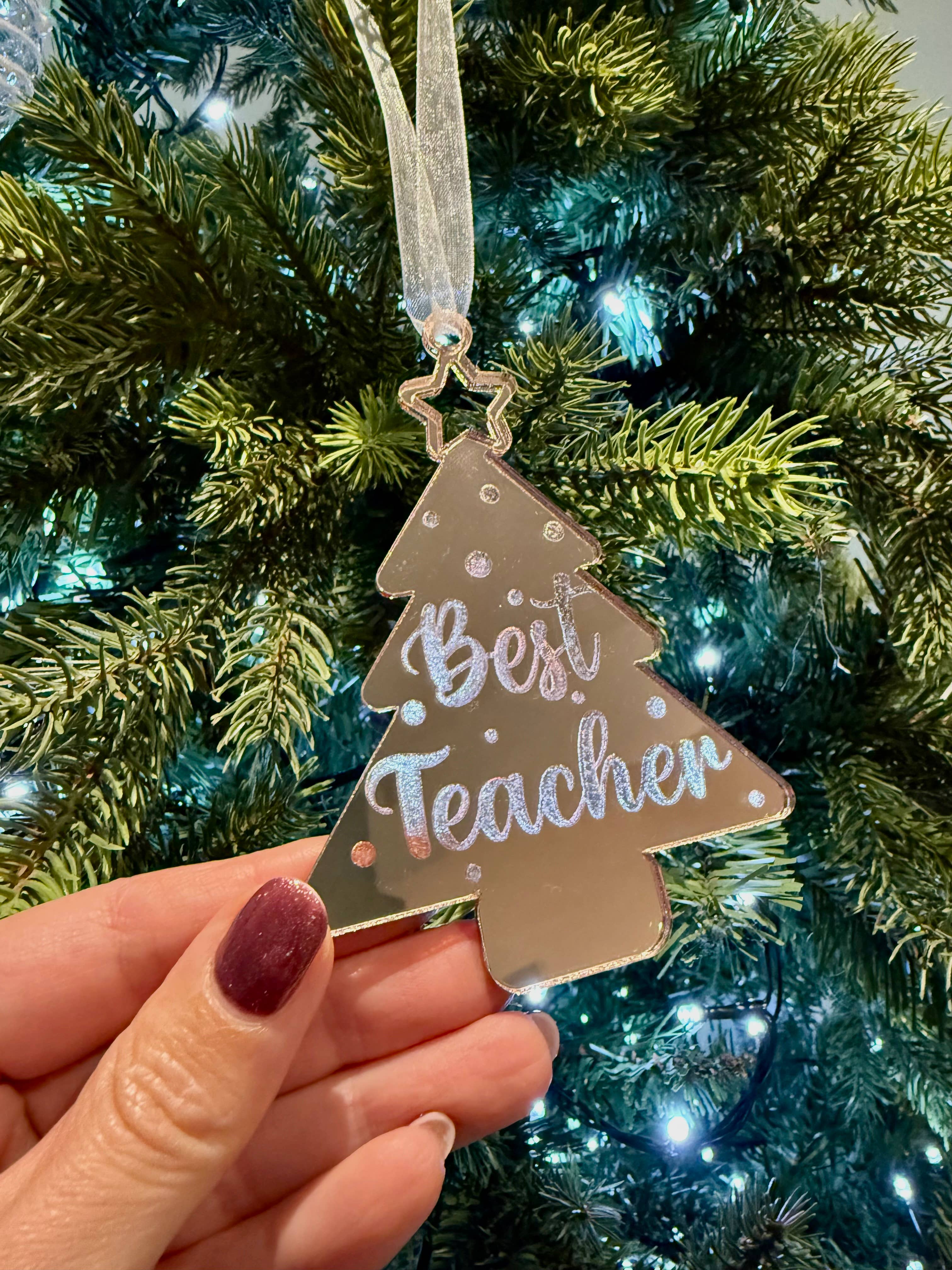 Decorative ornament with 'Best Teacher' text held in front of a Christmas tree.